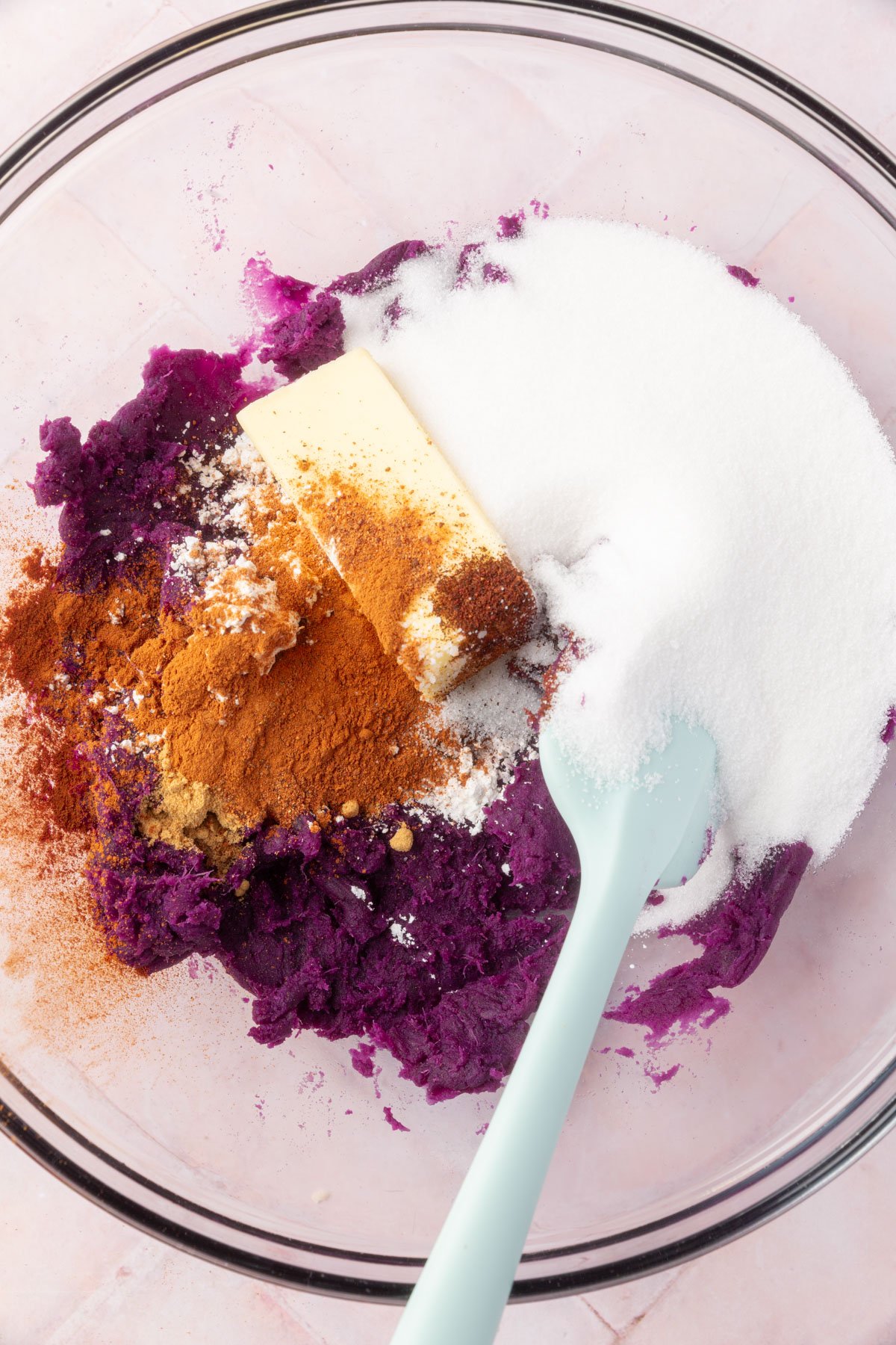 A bowl with mashed purple sweet potato, butter, granulated sugar, and spices like cinnamon and ginger, ready to be mixed for the pie filling.