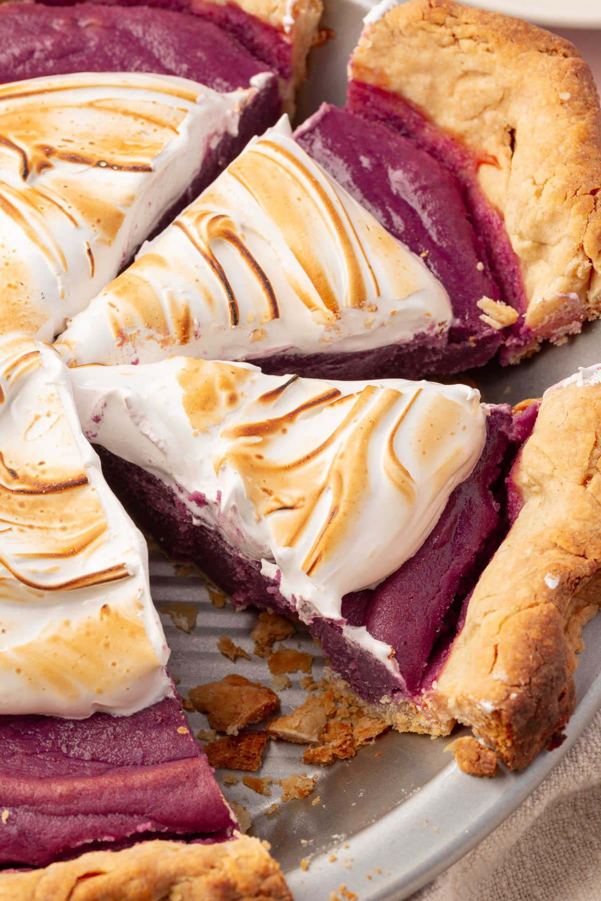 Close-up of a gluten-free purple sweet potato pie with a toasted marshmallow meringue topping, showing sliced portions and flaky crust, with crumbs scattered in the pie tin.