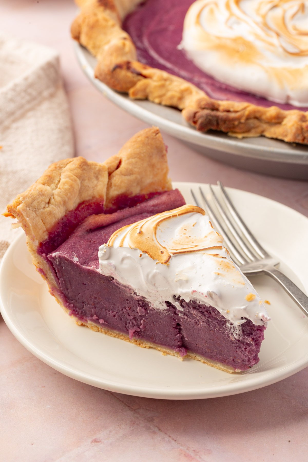 A close-up of a slice of purple sweet potato pie, focusing on the smooth purple filling, golden meringue, and flaky gluten-free crust with the rest of the gluten-free pie in the background.