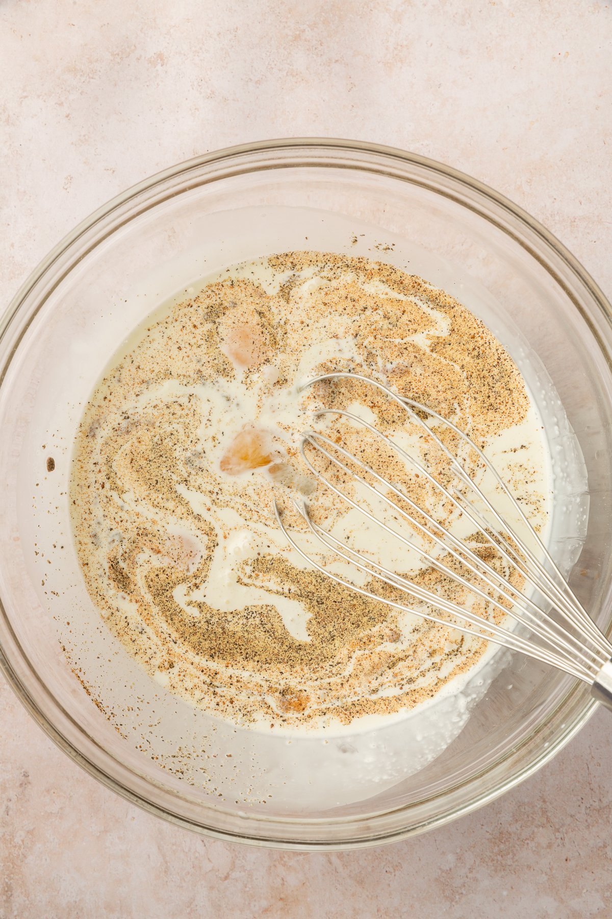Image of heavy cream, milk, salt, pepper and nutmeg whisked together in a clear mixing bowl.