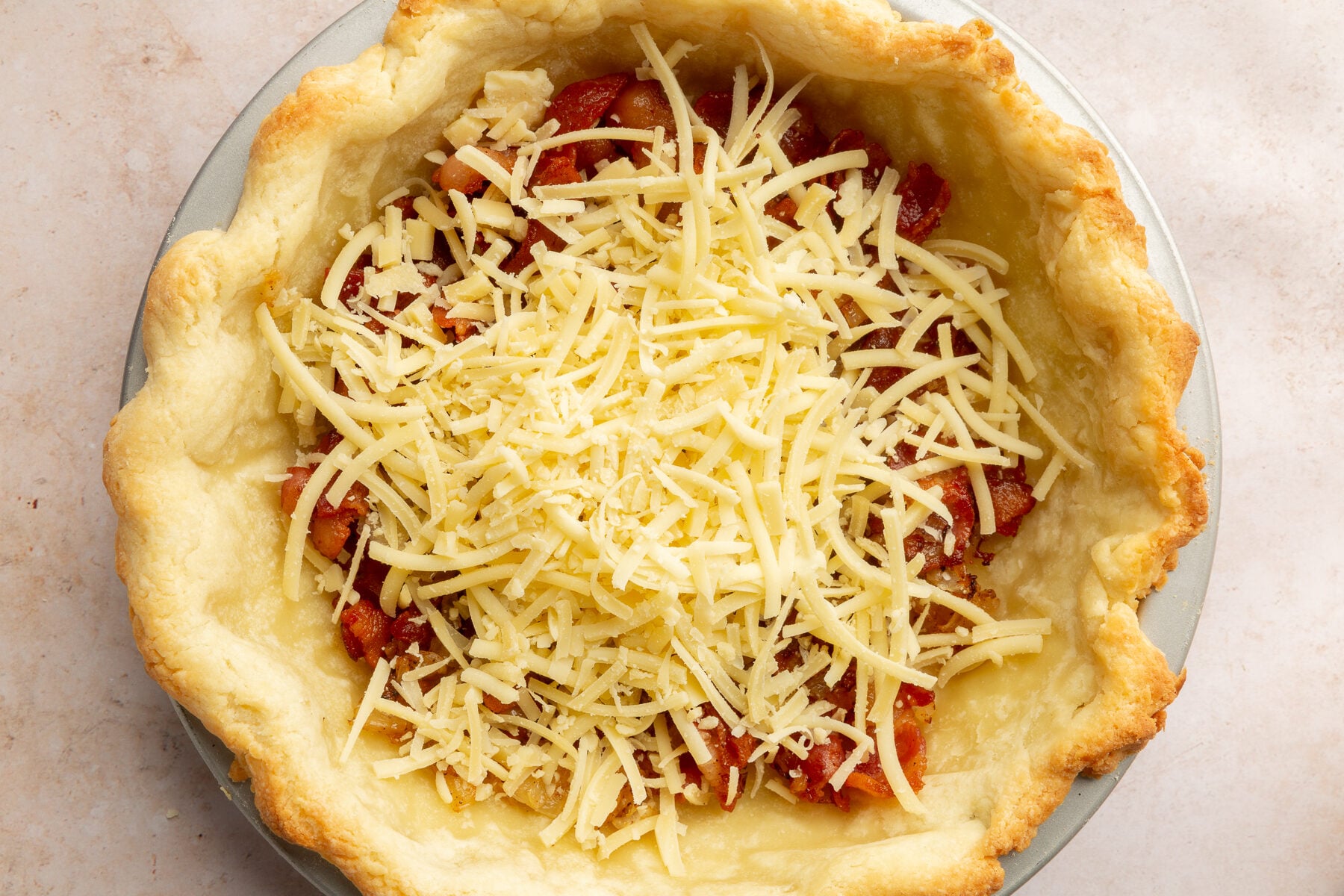 Image of gluten-free pie crust, cooked bacon, and gruyère cheese in a pie dish.