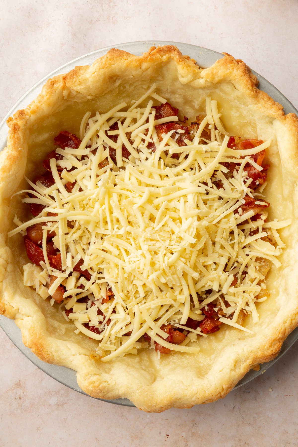 Image of gluten-free pie crust, cooked bacon, and gruyère cheese in a pie dish.
