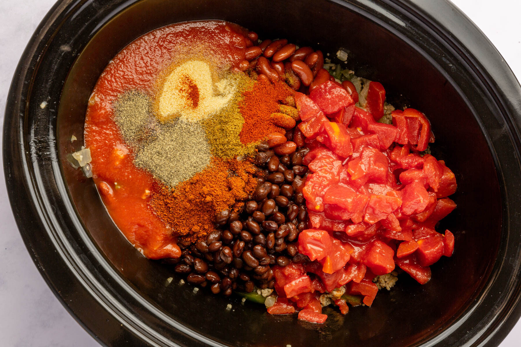 Crock pot filled with unmixed ingredients of diced tomatoes, black beans, kidney beans, cooked ground turkey, tomato sauce, paprika, cumin, salt, pepper, onion powder and chili powder.