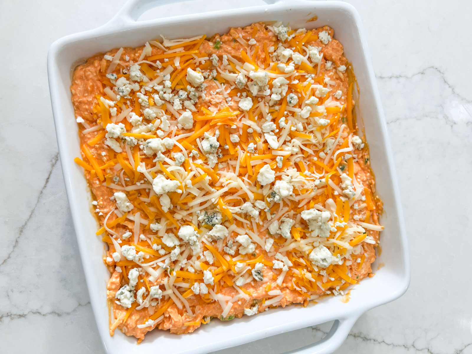 Unbaked buffalo chicken dip spread in a white baking dish, topped with shredded cheese and crumbled blue cheese.