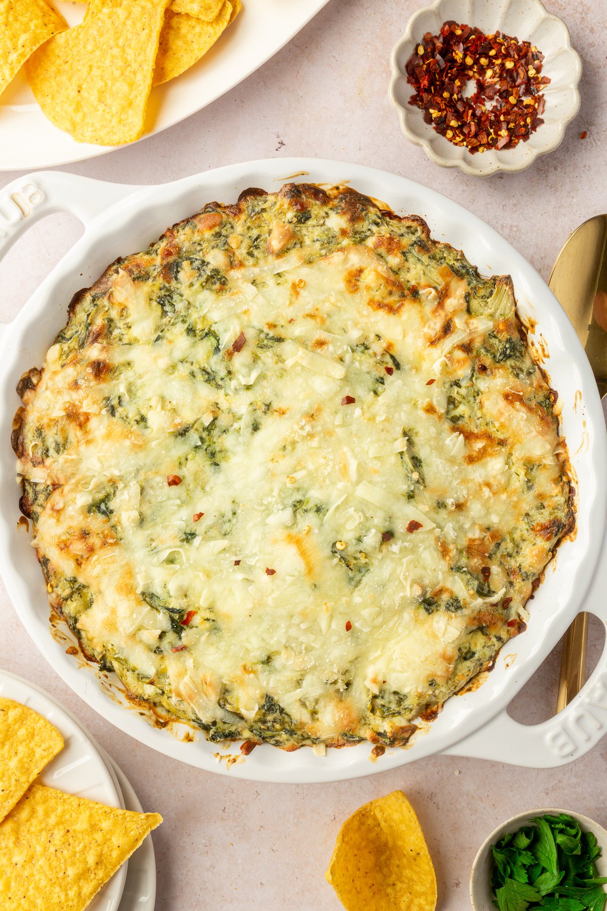 Finished spinach artichoke dip in a white pie dish sitting on a tabletop with chips and other garnished surrounding it.