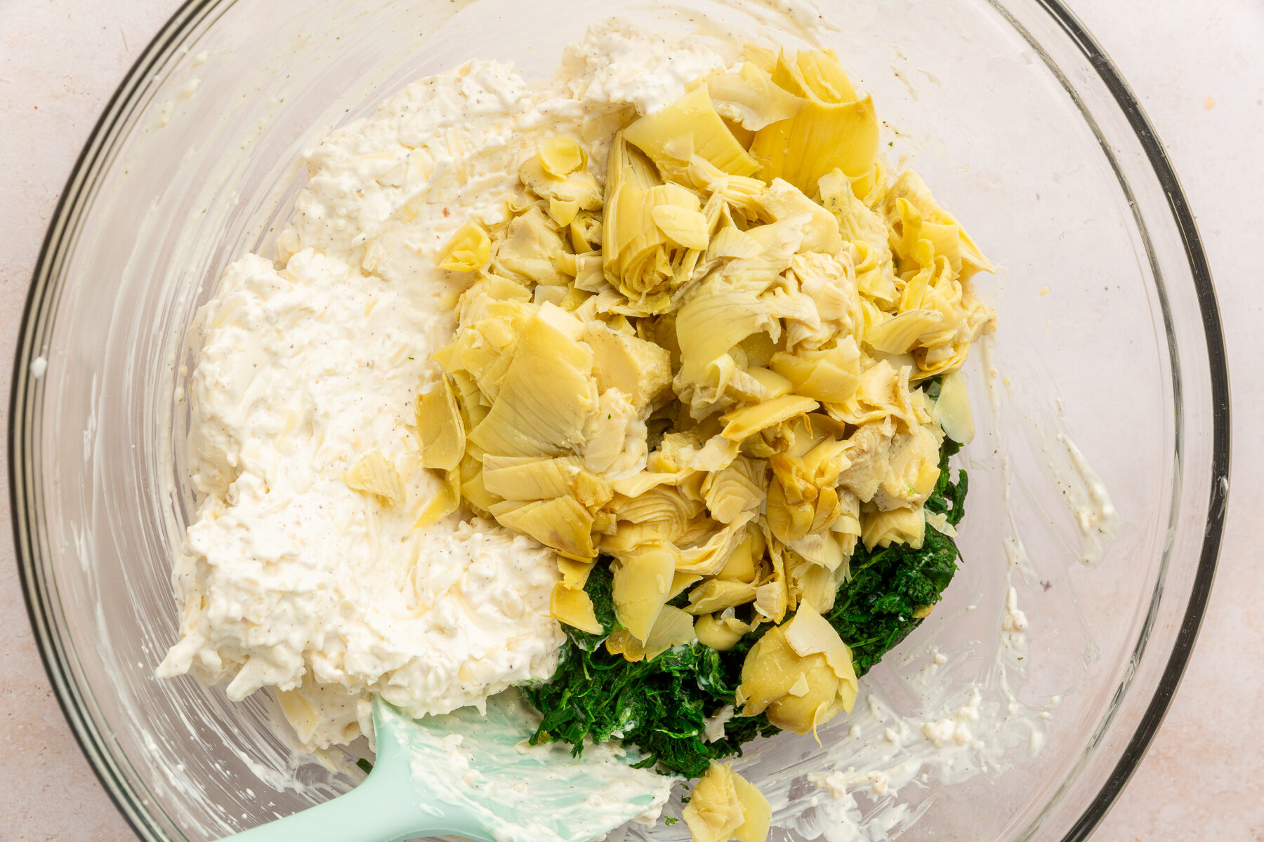 Ingredients for spinach artichoke dip including spinach, artichoke, sour cream, cream cheese, garlic, salt, pepper, mozzarella and Parmesan cheese together in a large, clear mixing bowl, not yet mixed together.