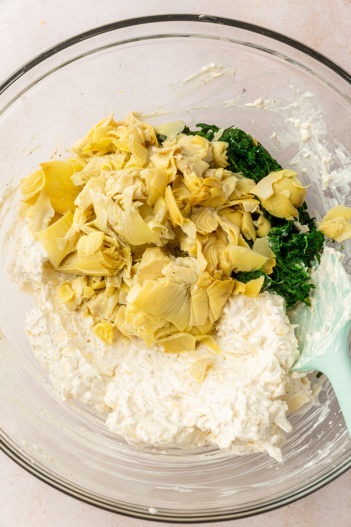 Ingredients for spinach artichoke dip including spinach, artichoke, sour cream, cream cheese, garlic, salt, pepper, mozzarella and Parmesan cheese together in a large, clear mixing bowl, not yet mixed together.