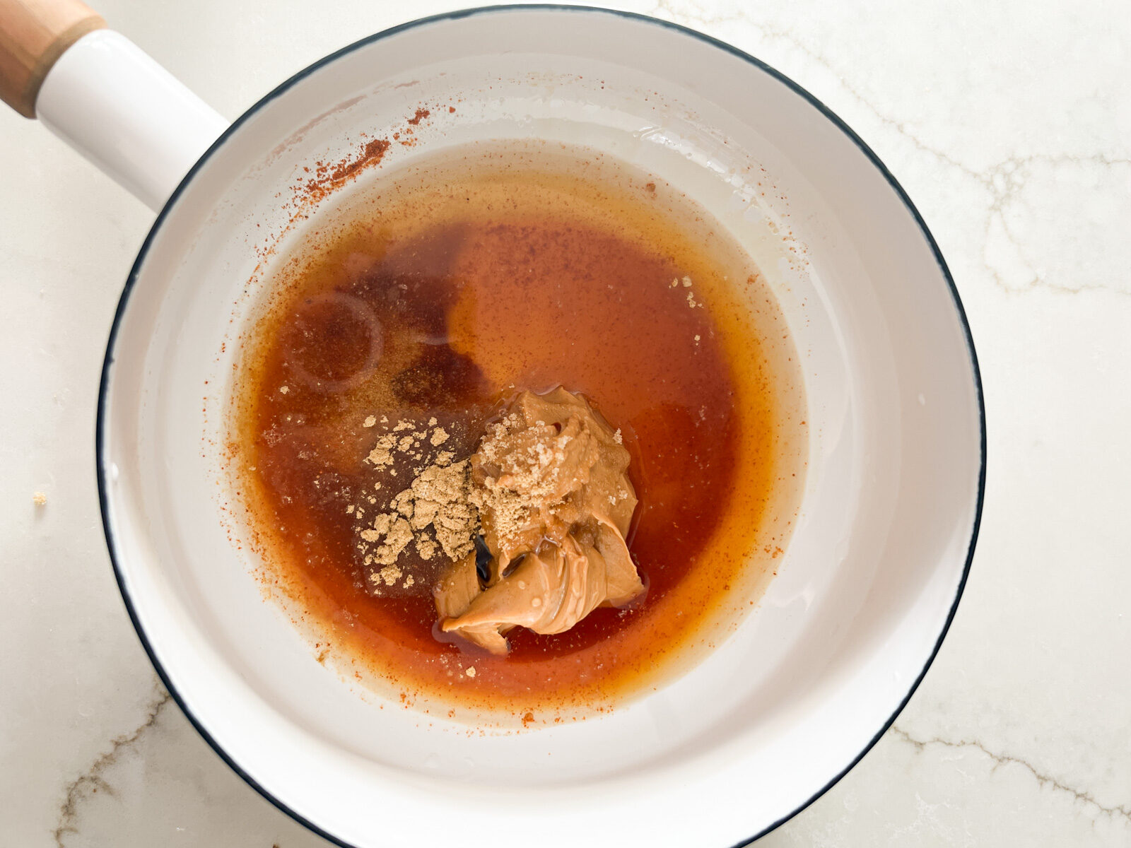 A white saucepan with peanut butter, brown sugar, ginger, cayenne pepper, water, tamari and lemon juice before mixing together.