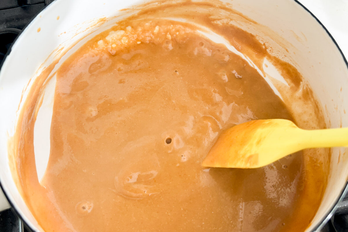 Thick peanut butter sauce cooking in a white saucepan on the stove top with a rubber spatula.