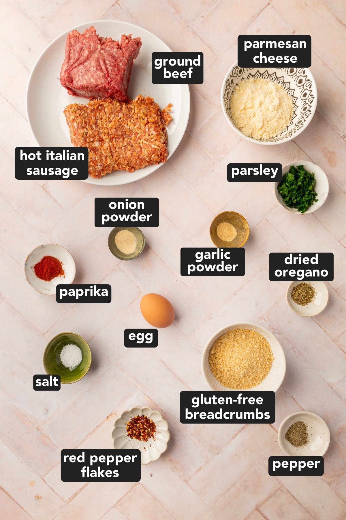Image of ground beef, Italian sausage, gluten-free breadcrumbs, an egg, parmesan cheese, fresh parsley, dried oregano, garlic powder, onion powder, paprika, salt, ground black pepper, and red pepper flakes, each in their own bowls resting on a brick countertop.