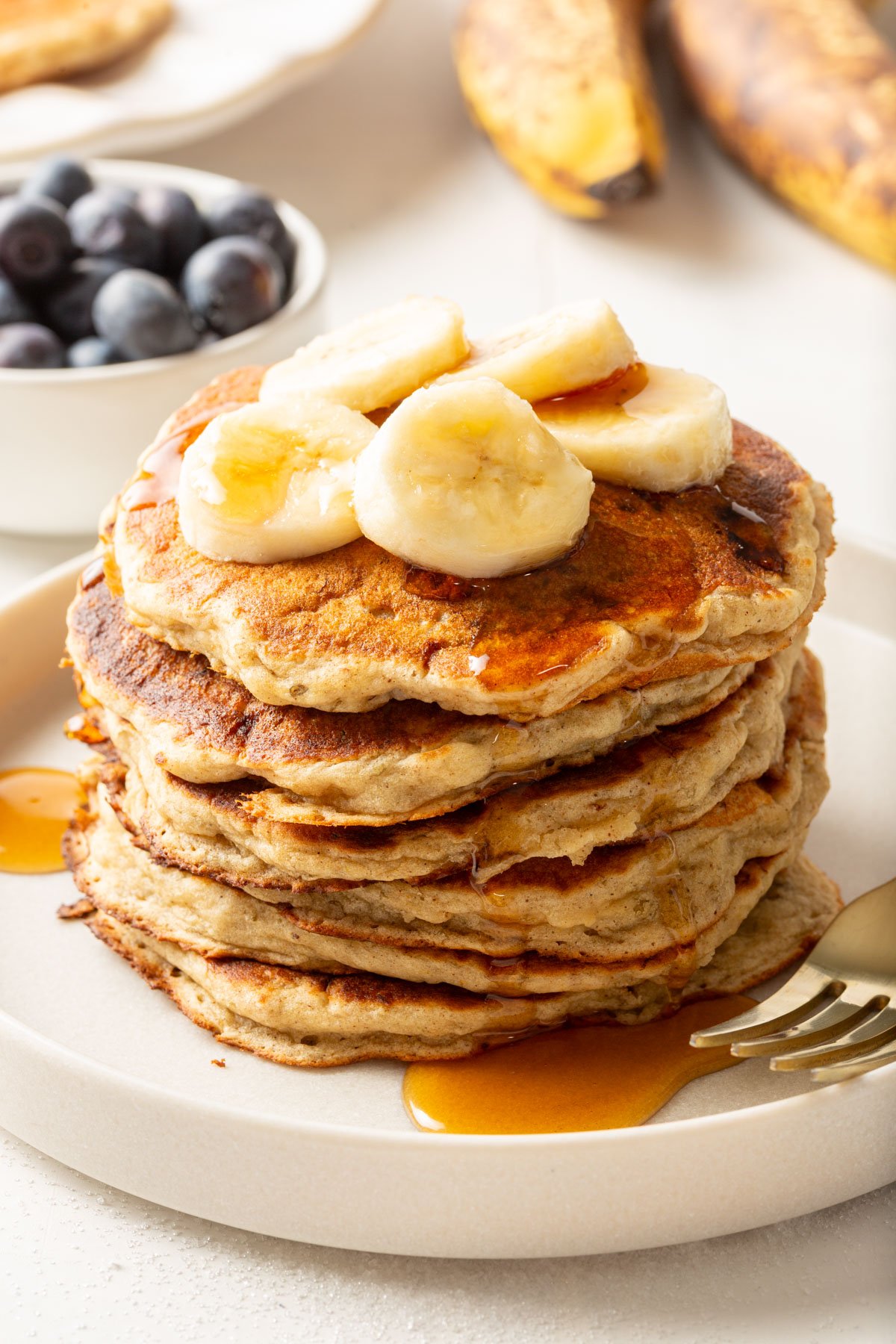 Stack of gluten-free banana pancakes with syrup and banana slices, with ripe bananas and a bowl of blueberries in the background.
