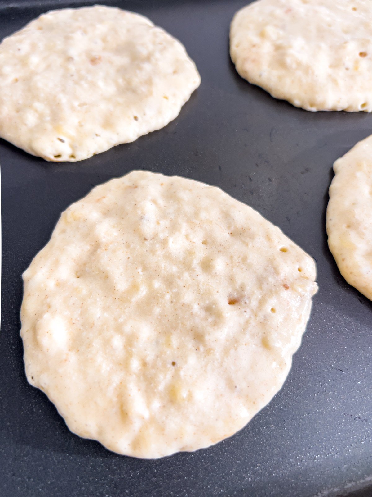 Close-up of gluten-free banana pancakes cooking on a griddle, showing a slightly uneven texture with air pockets.