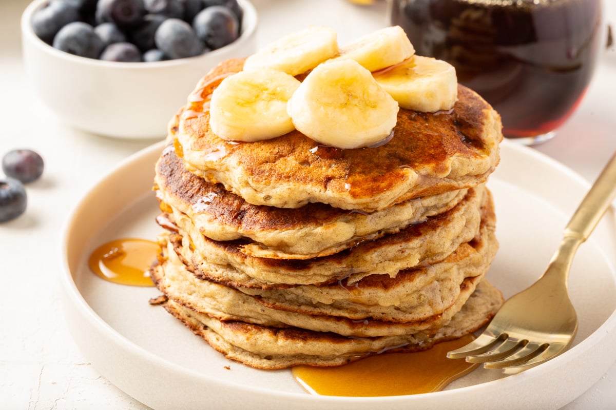 Stack of gluten-free banana pancakes on a white plate, topped with banana slices and drizzled with syrup, with a bowl of blueberries in the background.