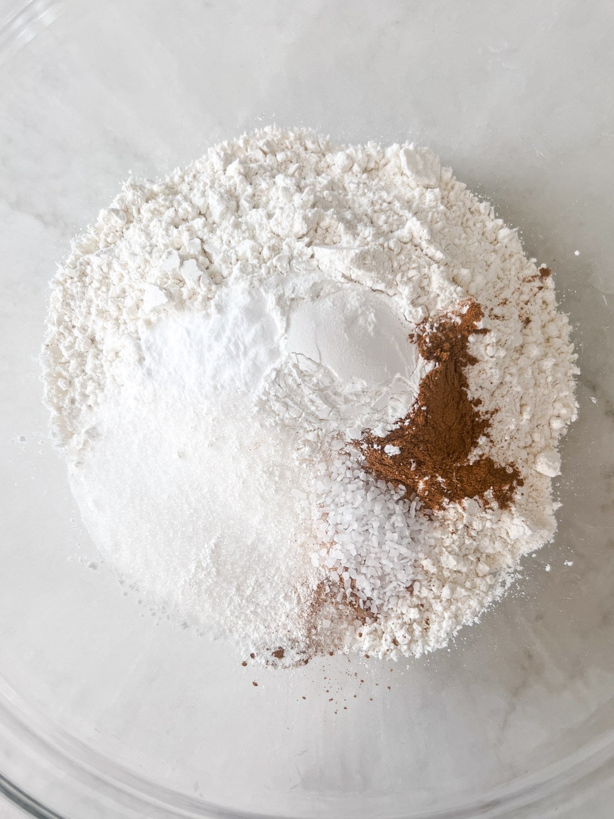 A glass mixing bowl containing dry ingredients for gluten-free banana pancakes, including flour, sugar, baking powder, salt, and cinnamon.
