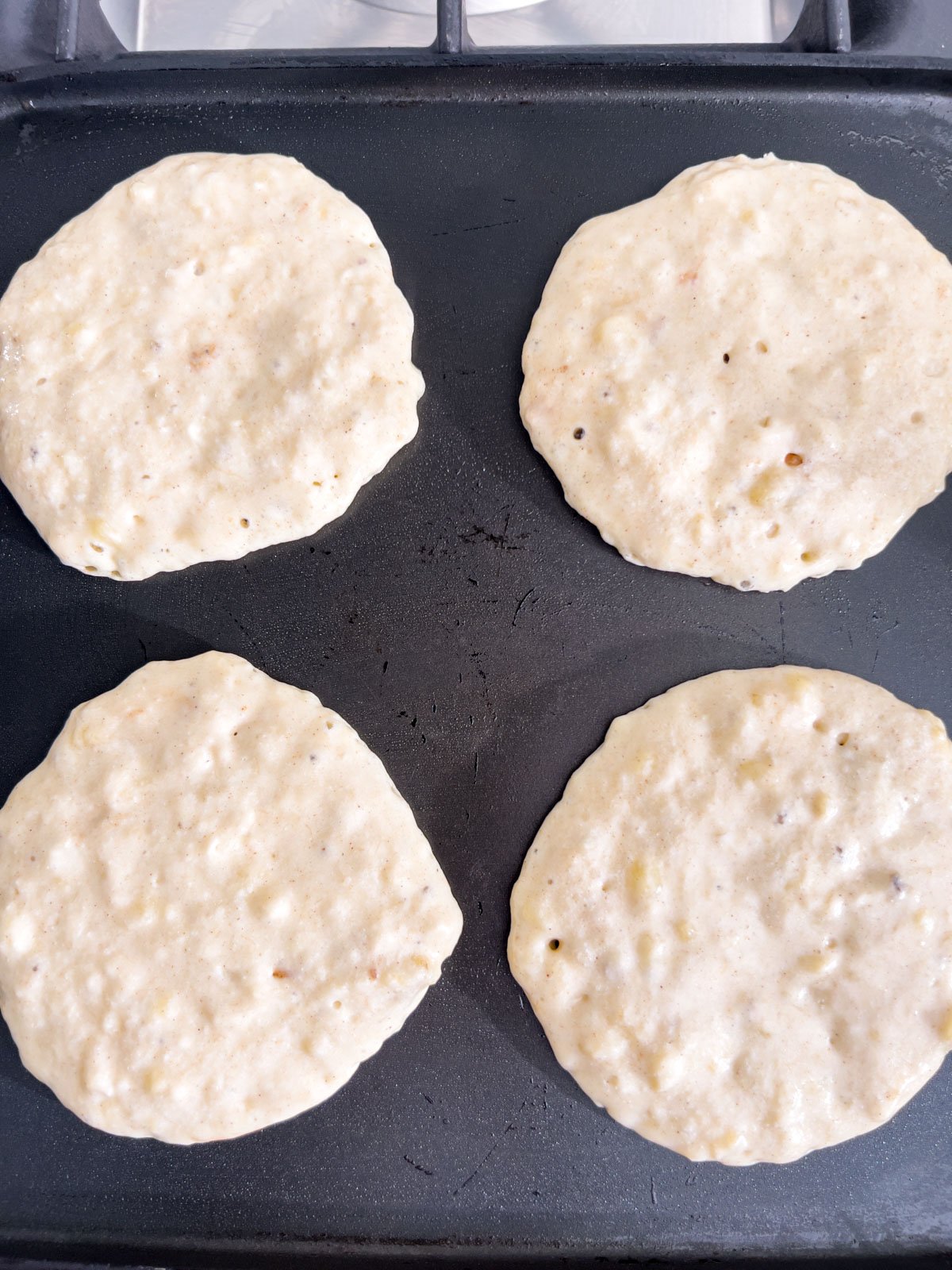 Four gluten-free banana pancakes cooking on a griddle, with bubbles starting to form on the surface.