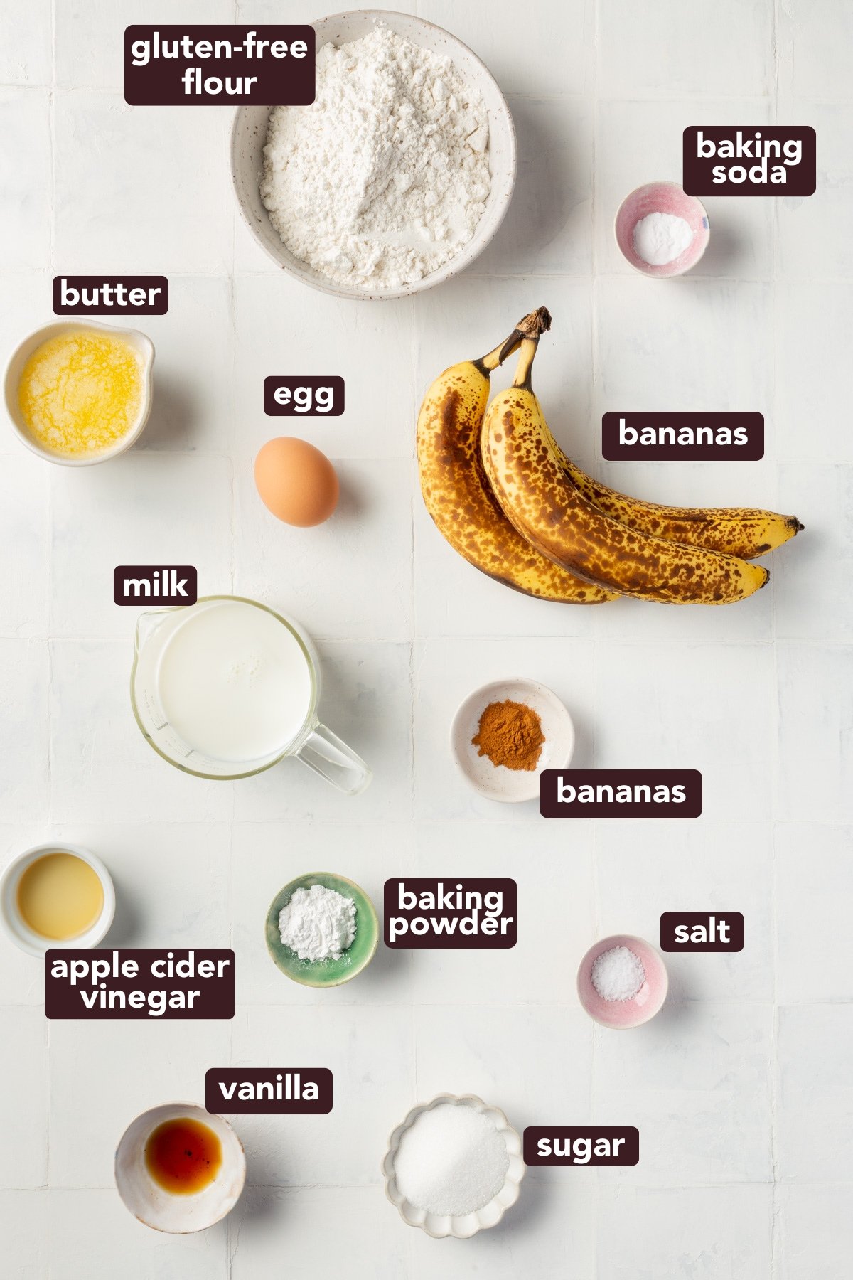 Flat lay of gluten-free banana pancake ingredients arranged on a white surface, including bananas, gluten-free flour, milk, egg, butter, sugar, baking powder, baking soda, salt, vanilla, cinnamon, and apple cider vinegar, each labeled with dark brown text.