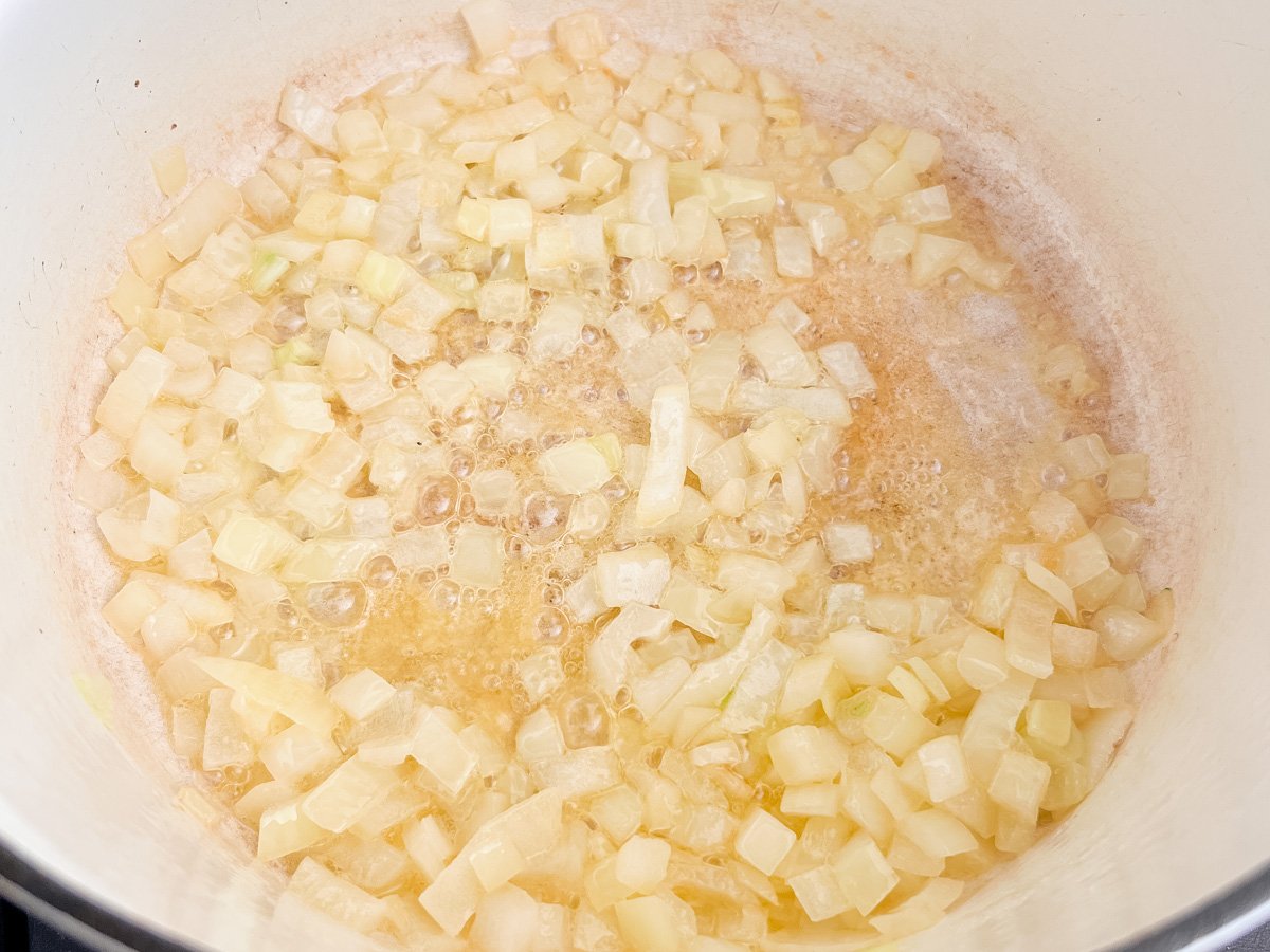 Sautéed diced onions sizzling in melted butter at the bottom of a white Dutch oven, starting the base of a broccoli cheddar soup.