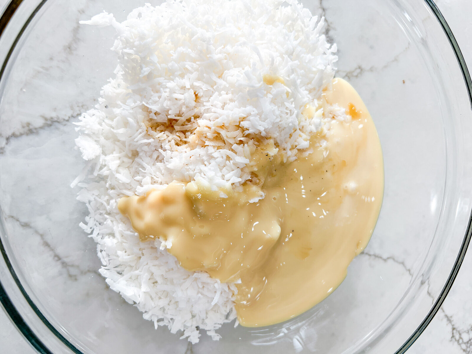 A glass mixing bowl containing shredded coconut, sweetened condensed milk, and vanilla extract before mixing.