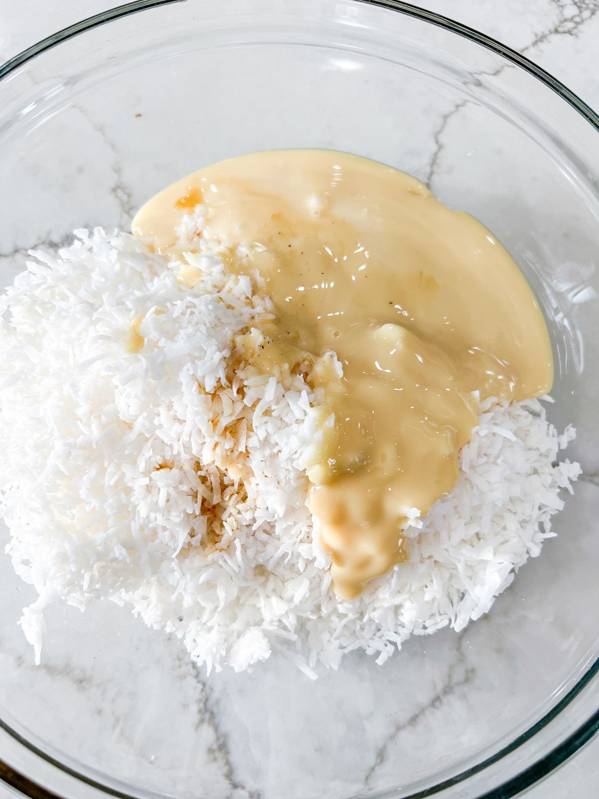 A glass mixing bowl containing shredded coconut, sweetened condensed milk, and vanilla extract before mixing.