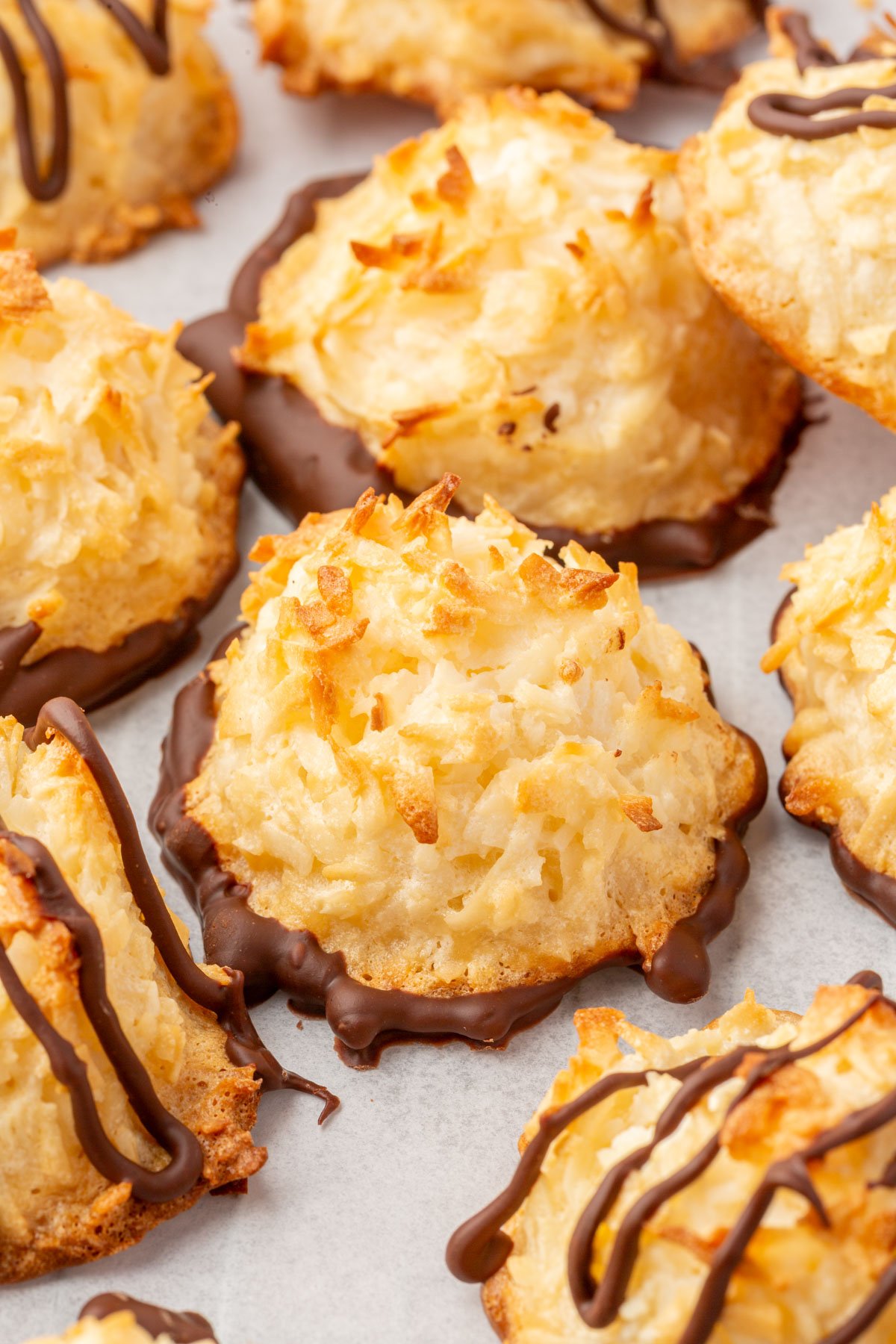 Several baked coconut macaroons with chocolate bottoms, some drizzled with additional chocolate, on parchment paper.