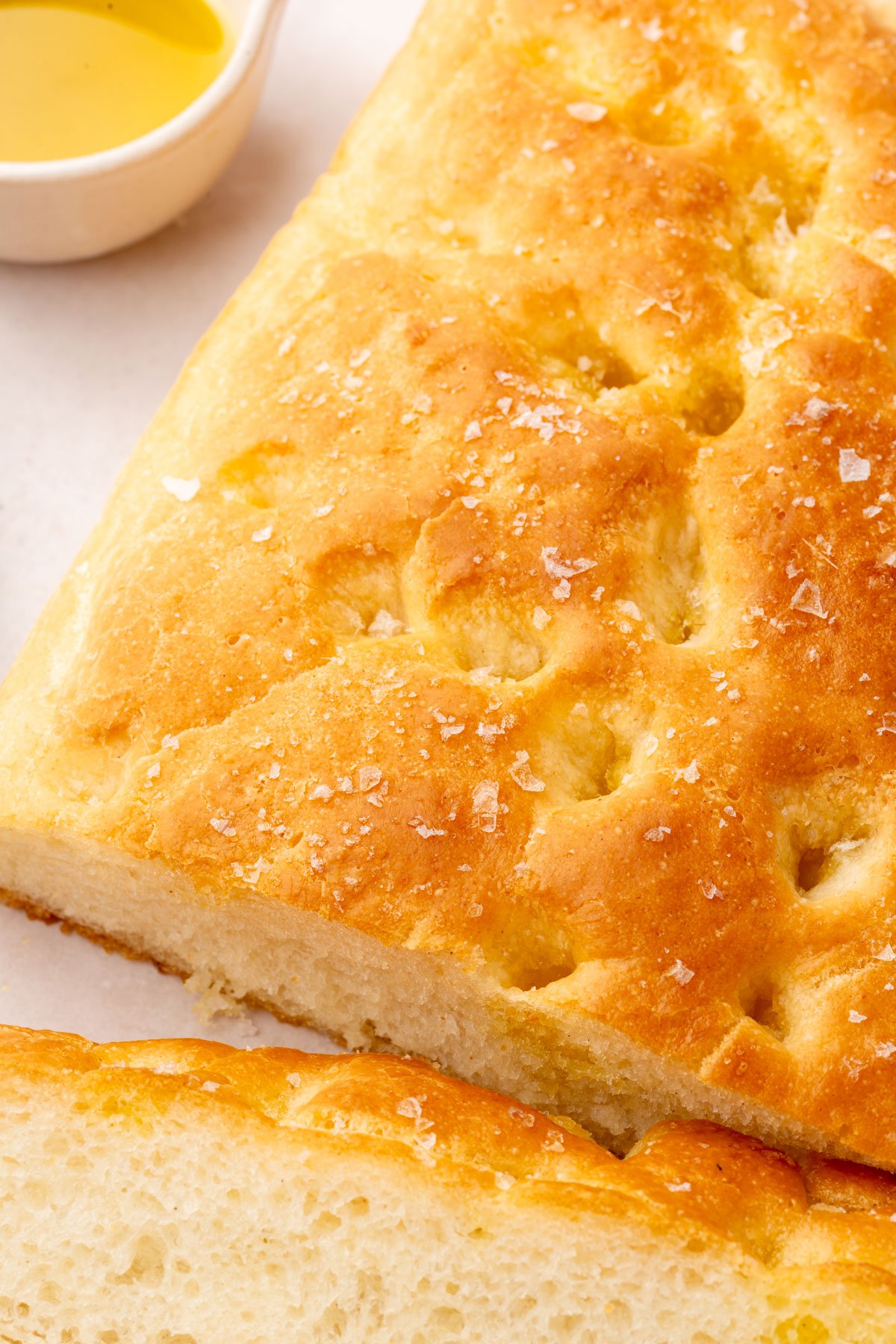 Close-up of golden-brown gluten-free focaccia showing a crispy, dimpled crust and sea salt flakes, with a small bowl of olive oil partially in frame.