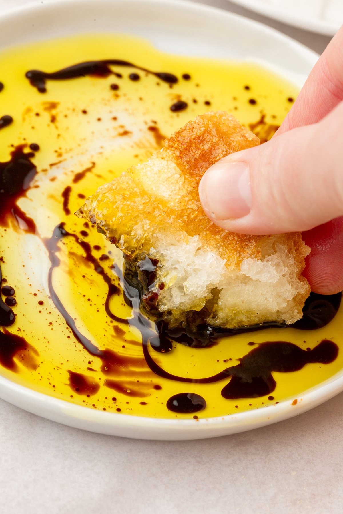Hand dipping a torn piece of gluten-free focaccia into a swirl of olive oil and balsamic vinegar on a small white plate.