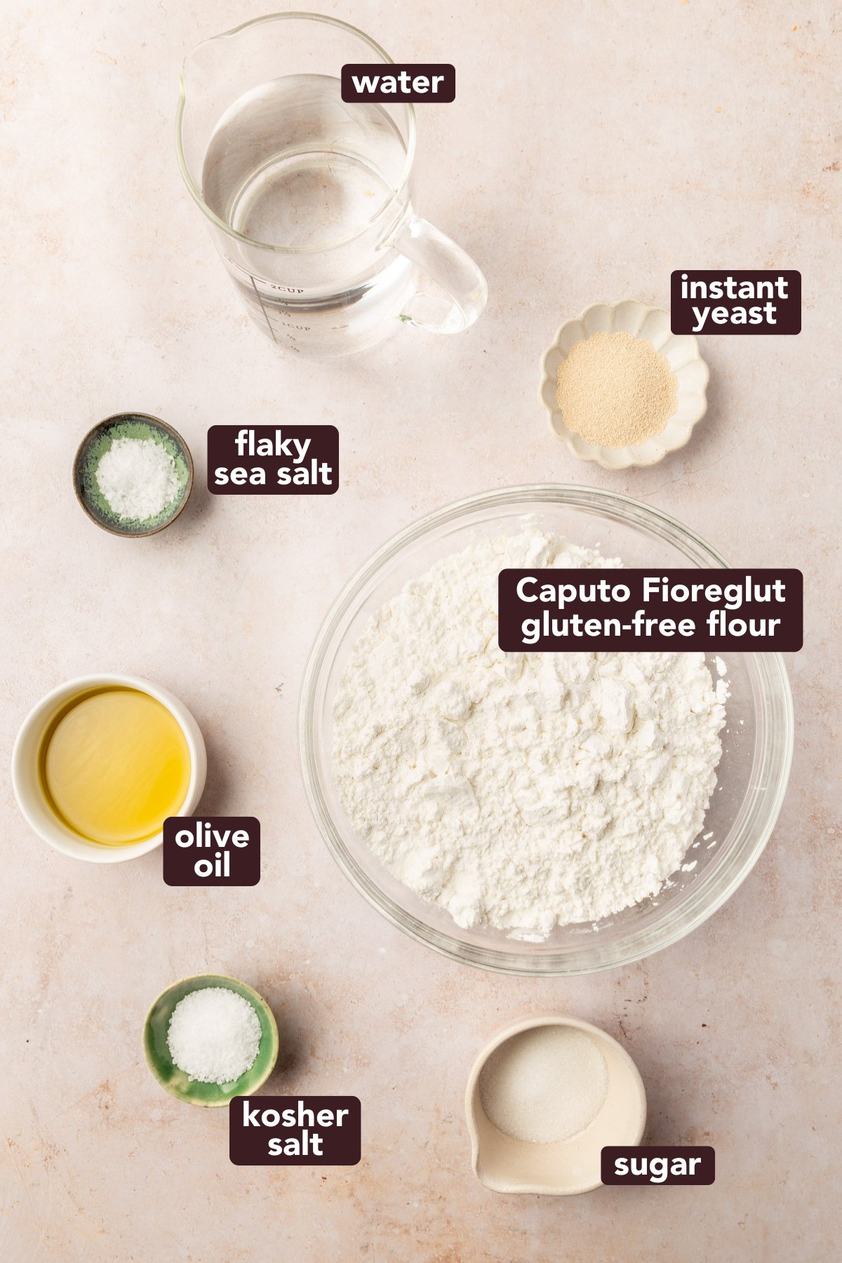 Overhead shot of ingredients for gluten-free focaccia, including Caputo Fioreglut flour, instant yeast, warm water, olive oil, sugar, kosher salt, and flaky sea salt.