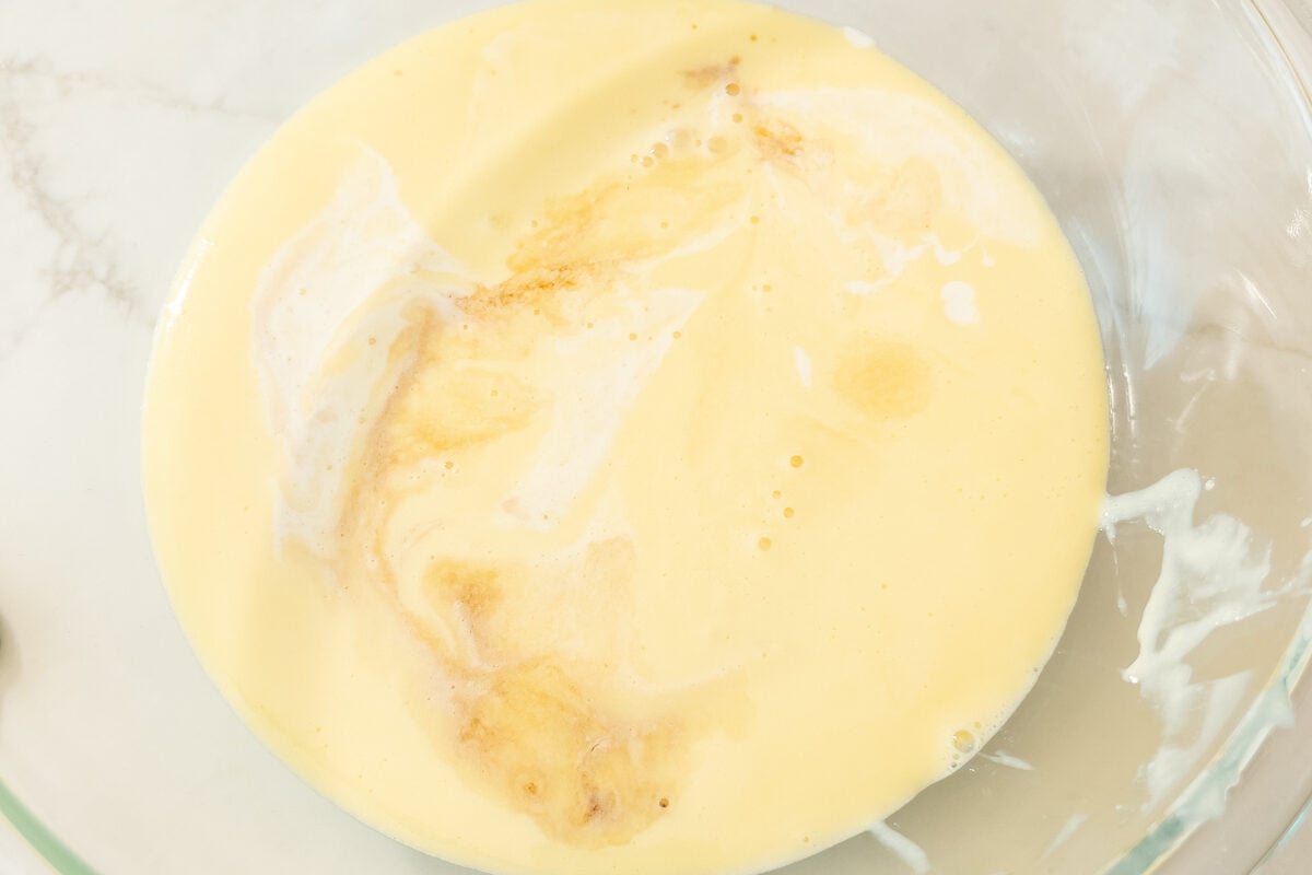 Top view of vanilla extract and cream being added to the cooled custard base in a mixing bowl.