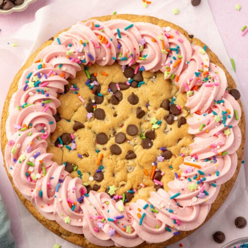 Gluten-free cookie cake decorated with pink buttercream swirls and colorful sprinkles, styled on parchment with chocolate chips and a dish towel nearby.