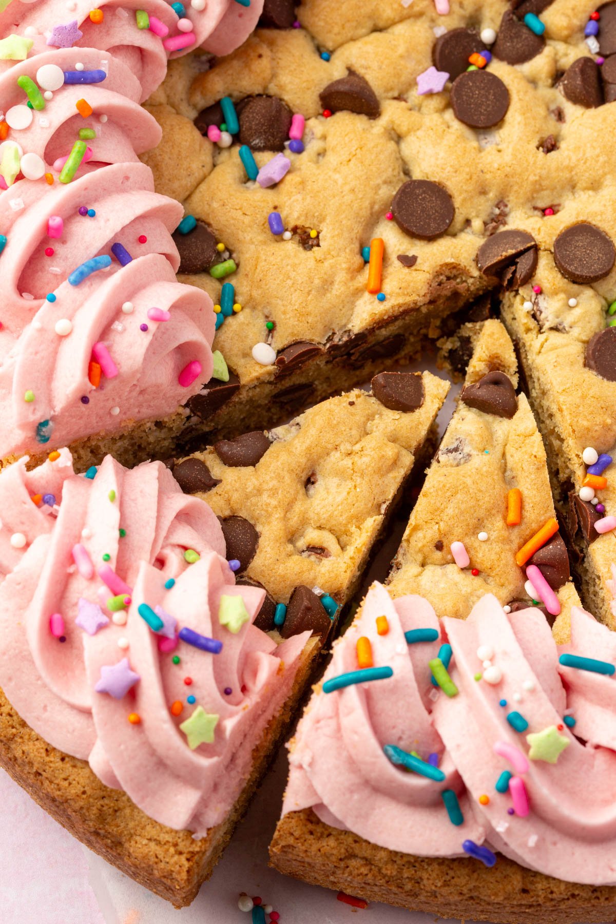 Close-up view of two slices cut from a gluten-free cookie cake, showing chocolate chips and pink piped frosting topped with sprinkles.
