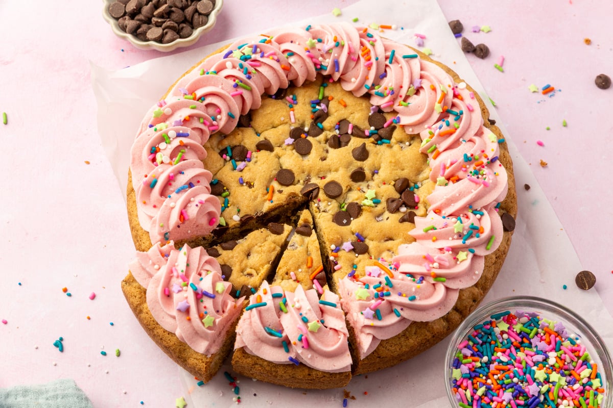 Angled view of a gluten-free cookie cake with two slices cut, showcasing pink frosting rosettes and rainbow sprinkles on a parchment-lined surface.