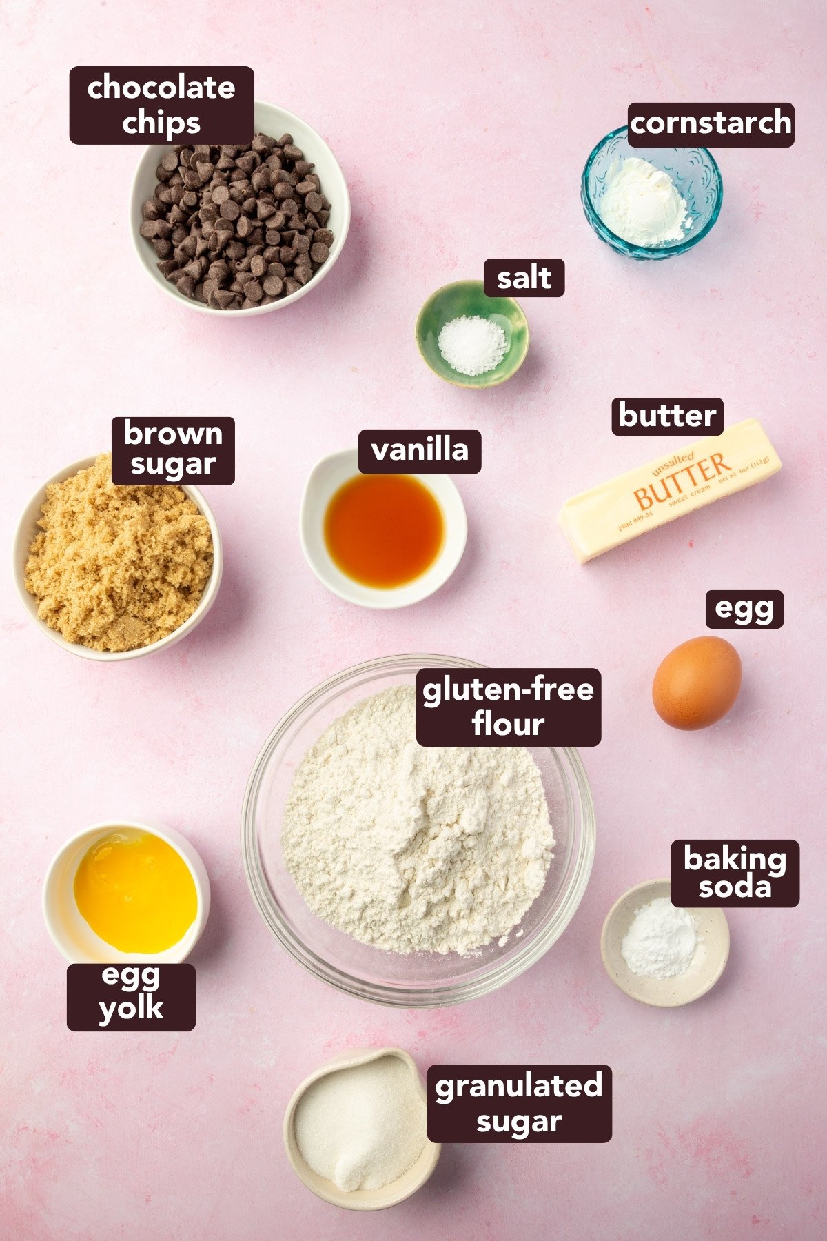 Overhead of labeled ingredients for a gluten-free cookie cake, including gluten-free flour, butter, sugars, vanilla, cornstarch, and chocolate chips on a pink background.