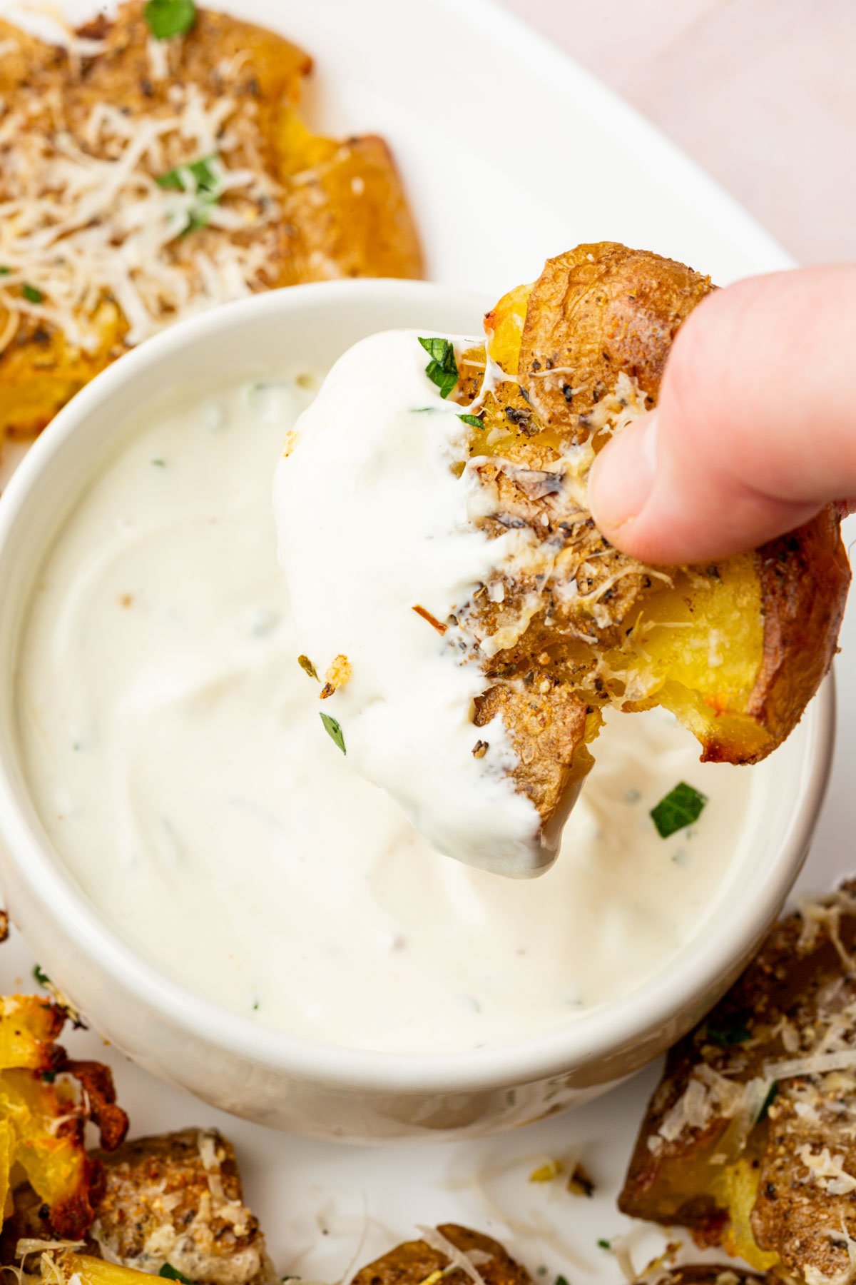 Hand dipping a cheesy smashed potato into a bowl of creamy white sauce.