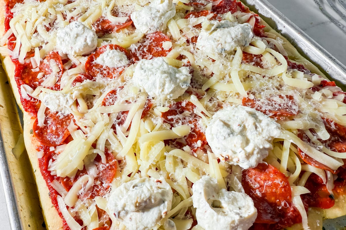 Parbaked focaccia pizza topped with sauce, cheese, pepperoni, and dollops of ricotta mixture, ready to bake.