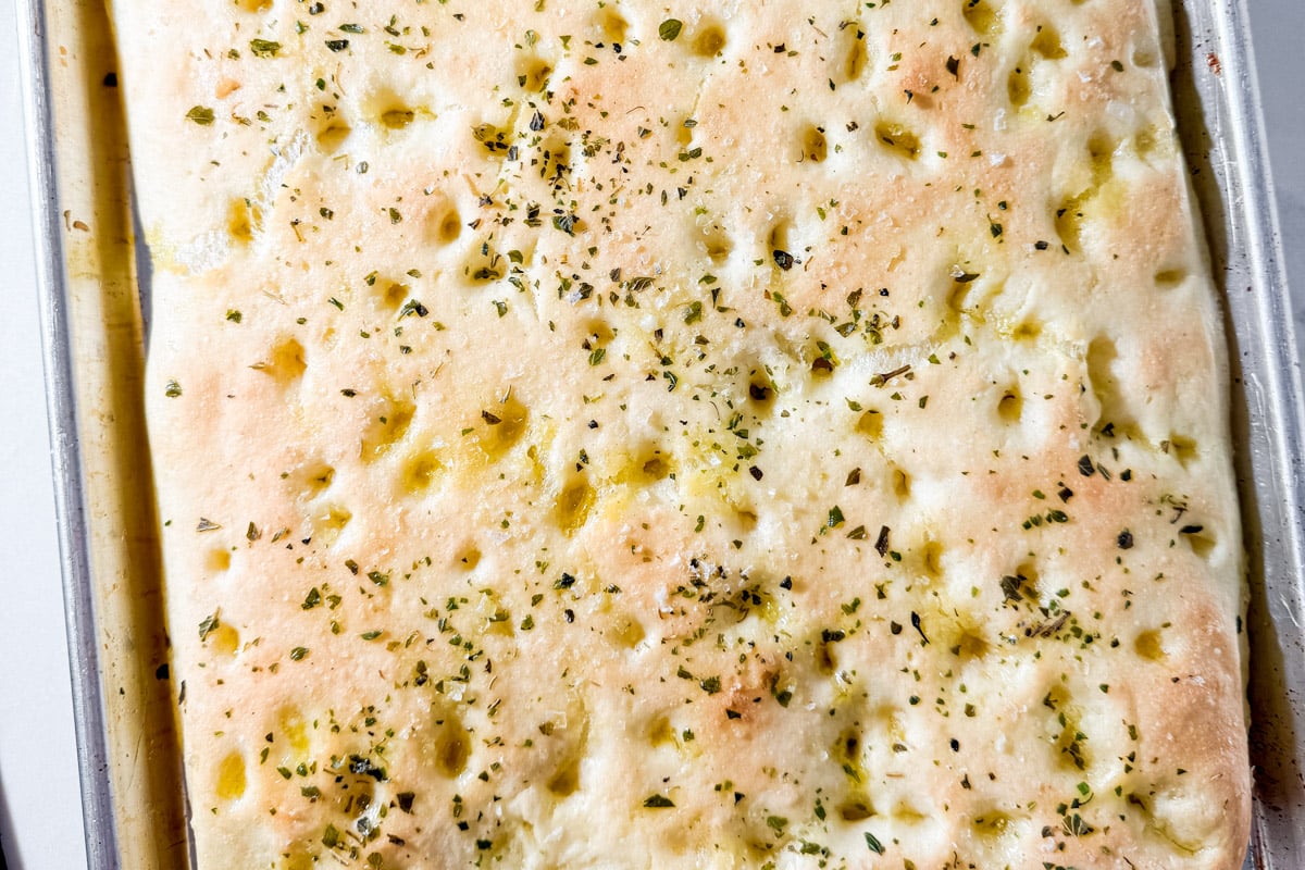 Lightly golden gluten-free focaccia crust after parbaking, topped with olive oil and herbs.