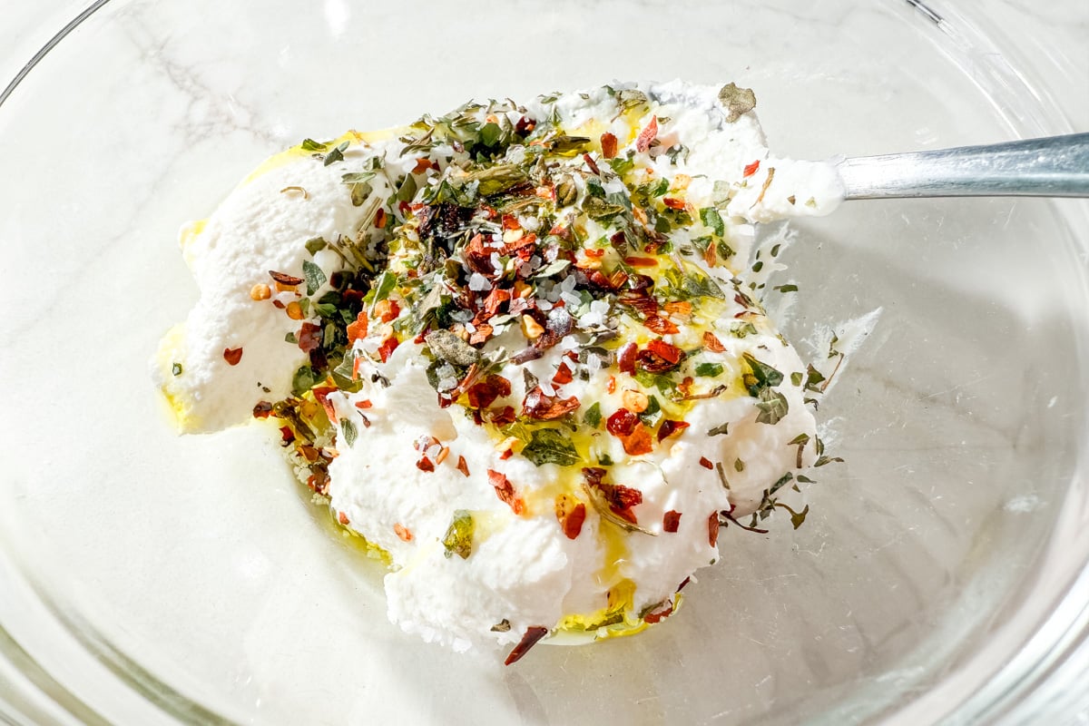 Bowl of ricotta topped with dried oregano, red pepper flakes, olive oil, and salt before mixing.
