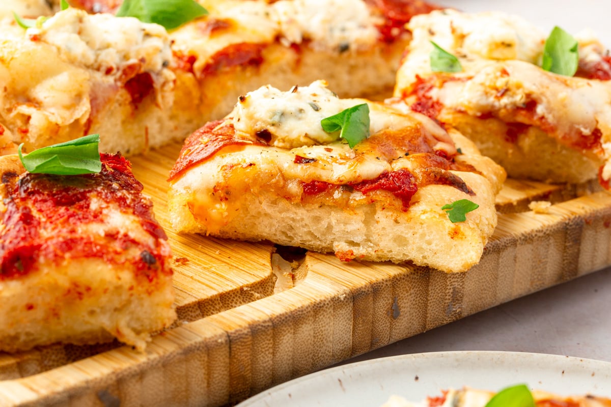 Sliced gluten-free focaccia pizza on a wood cutting board topped with basil and red pepper flakes.