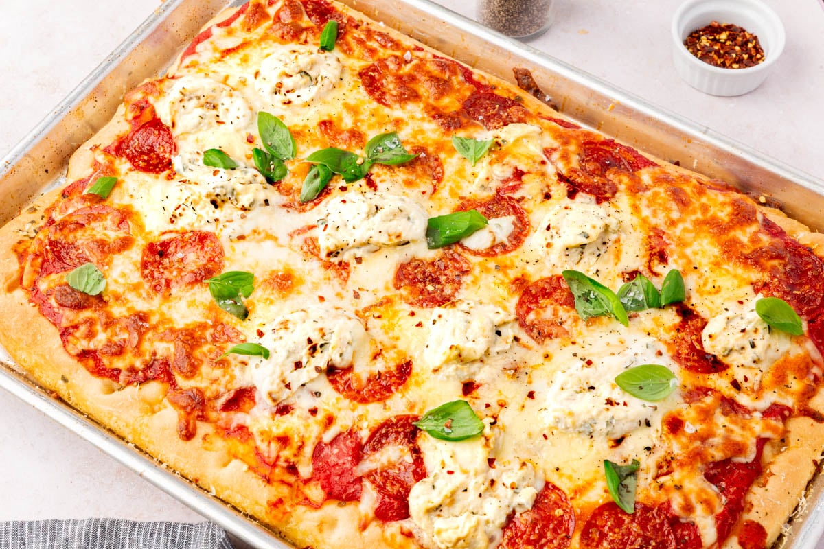 Fully baked gluten-free focaccia pizza topped with melted cheese, pepperoni, ricotta dollops, and fresh basil in a sheet pan.