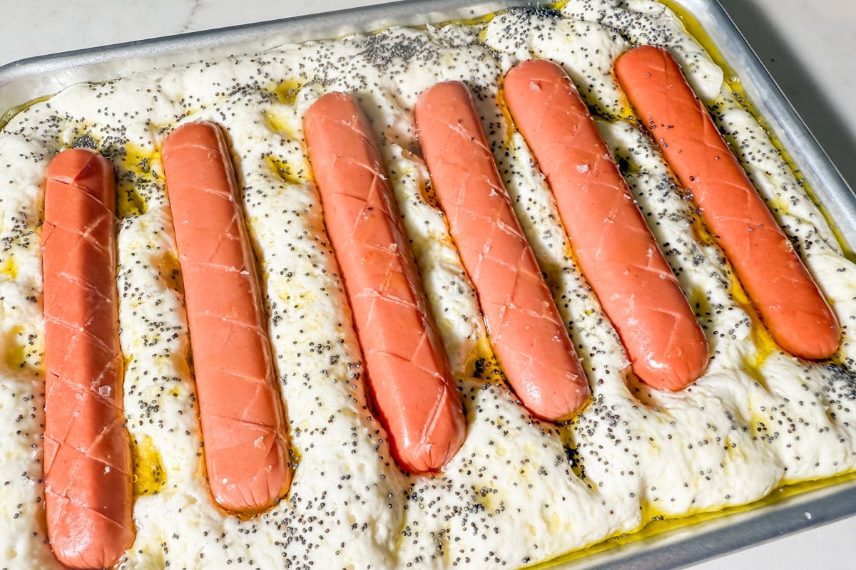 Unbaked gluten-free focaccia topped with scored hot dogs and poppy seeds, ready to go into the oven.