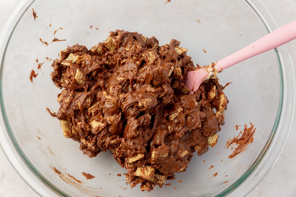 Chocolate-coated cereal mixture fully combined and clumped together in a glass bowl with a pink spatula.