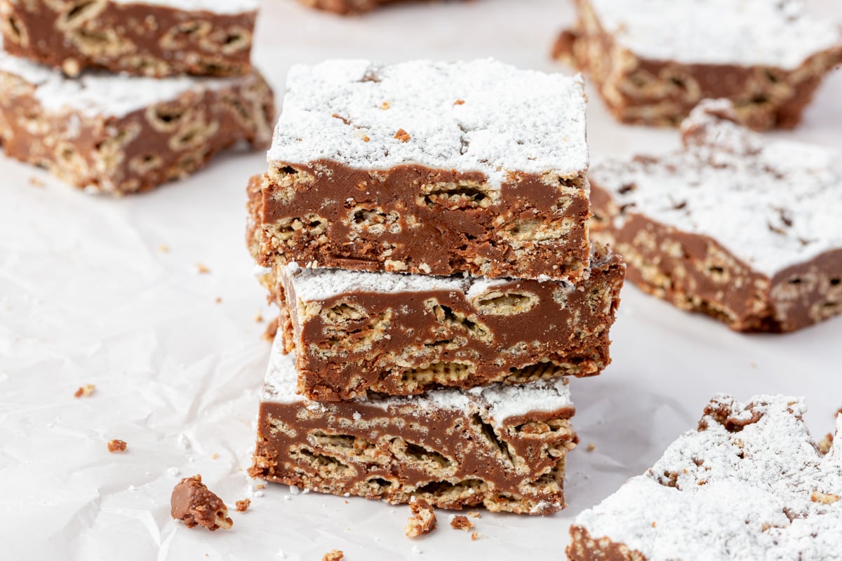 Three puppy chow bars stacked on parchment paper, with powdered sugar dusted on top and cereal pieces visible in the chocolate layer.