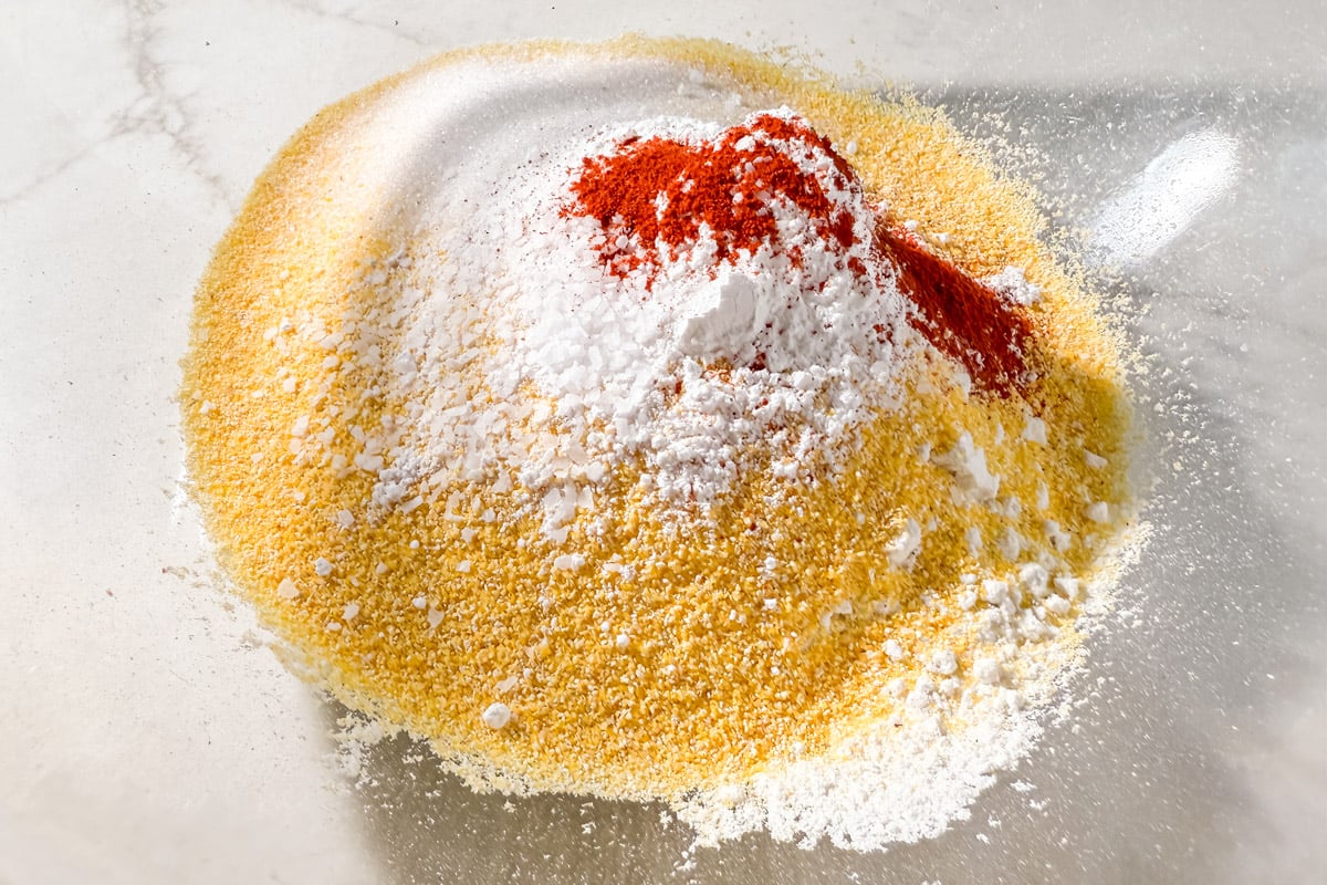 A pile of cornmeal, gluten-free flour, sugar, salt, and smoked paprika arranged in a mixing bowl before combining.