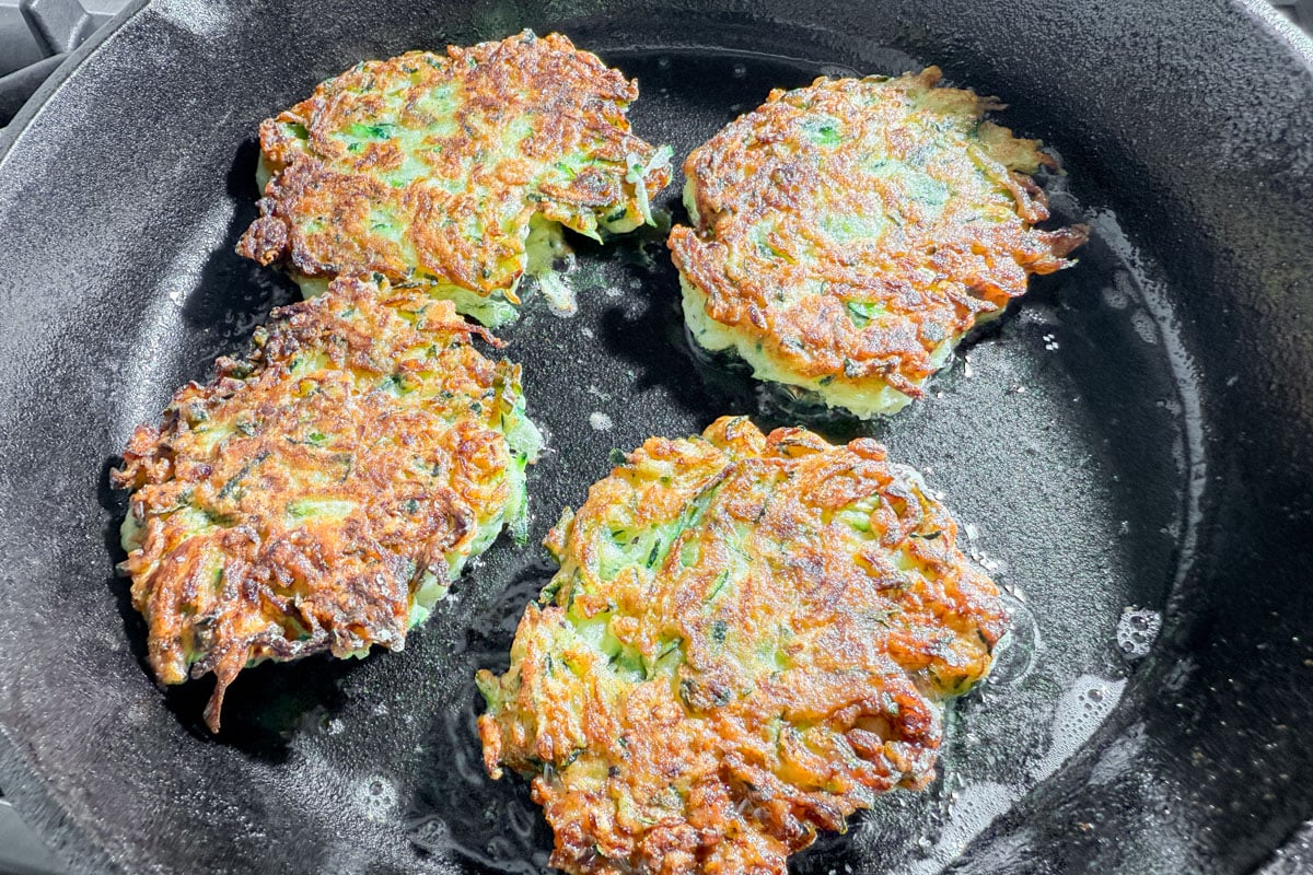 Three uncooked zucchini fritters sizzling in a cast iron pan before flipping.