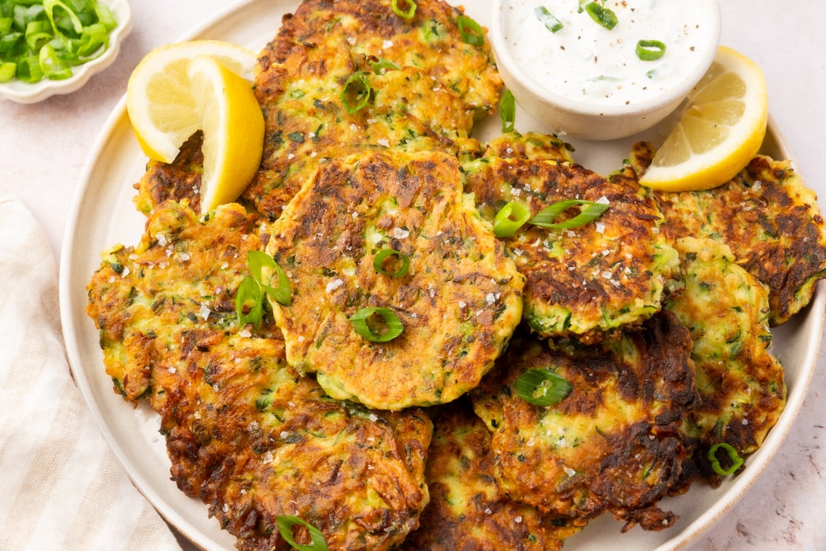 A plate full of crispy golden-brown zucchini fritters garnished with sliced green onions and lemon wedges, served with a bowl of sour cream dip.