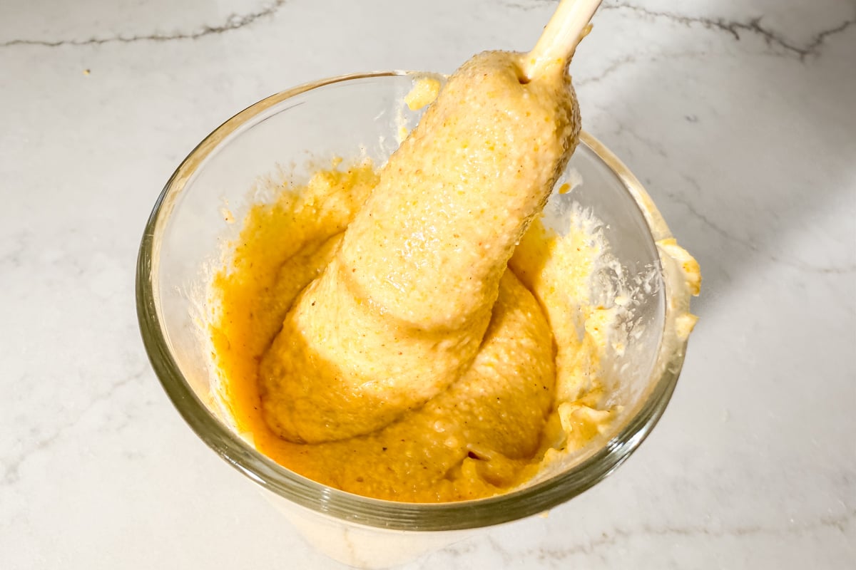 A skewered hot dog being dipped into a glass of thick gluten-free corn dog batter.