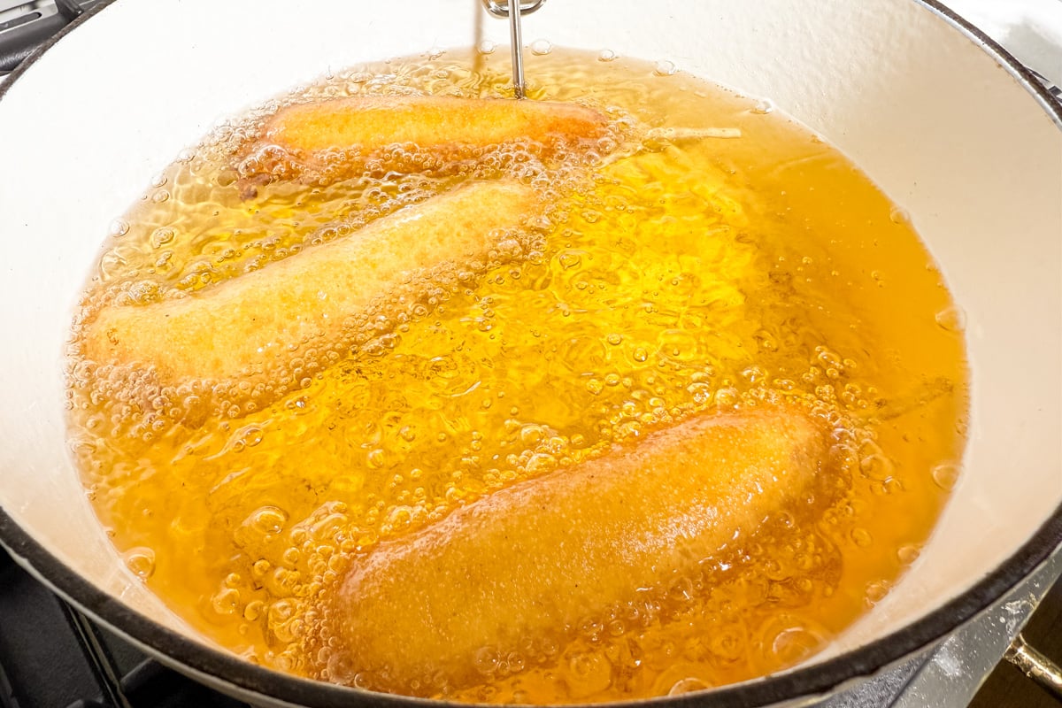 Three gluten-free corn dogs frying in bubbling oil inside a white Dutch oven.