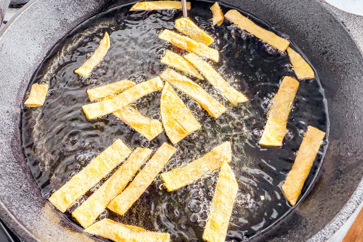 Corn tortilla strips frying in hot oil in a cast iron skillet.
