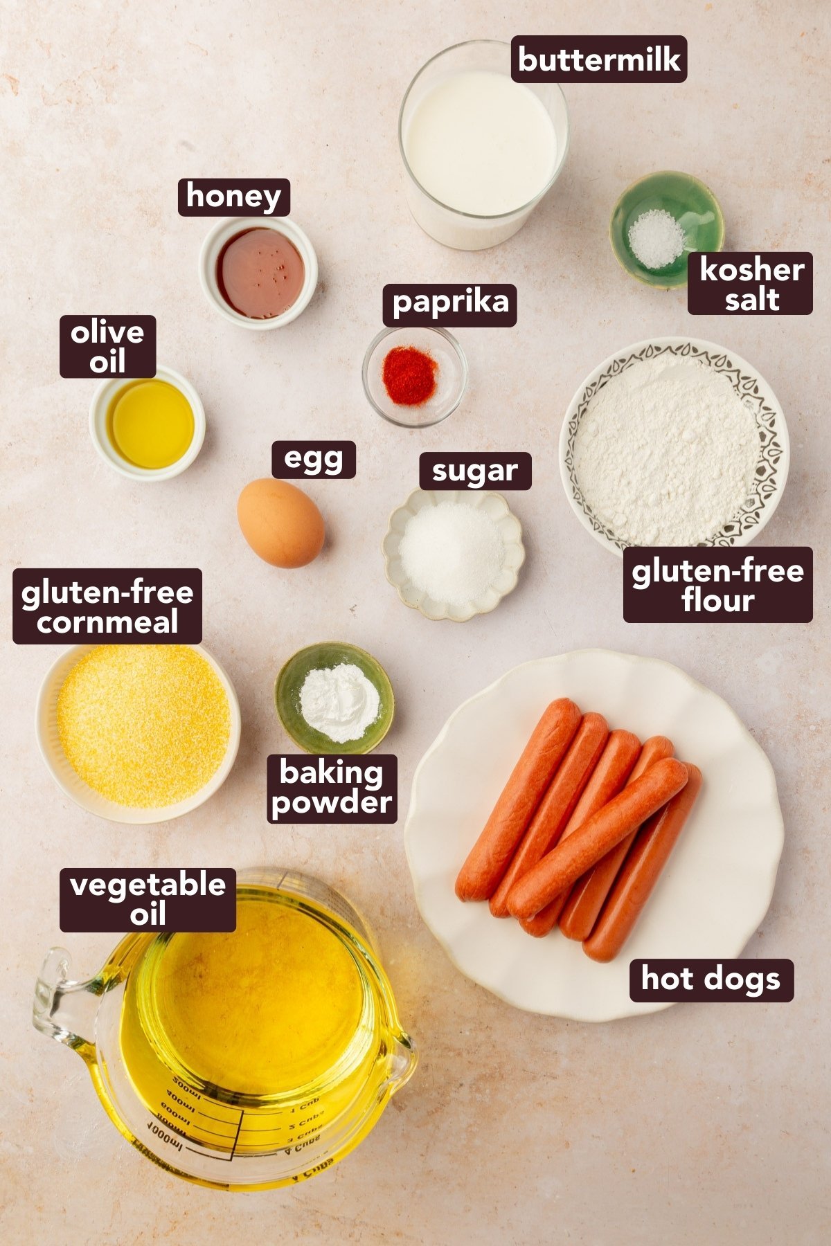 Overhead view of ingredients for gluten-free corn dogs including cornmeal, gluten-free flour, hot dogs, buttermilk, egg, honey, paprika, salt, sugar, olive oil, baking powder, and vegetable oil.