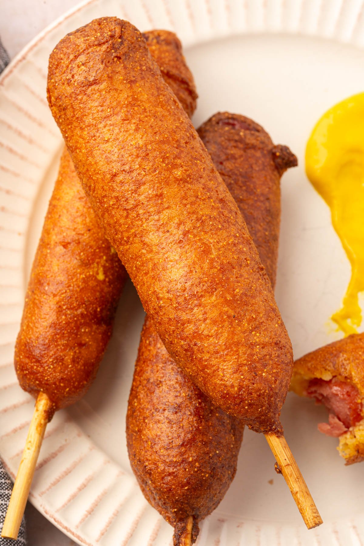Three gluten-free corn dogs on a white plate with a dollop of yellow mustard for dipping.