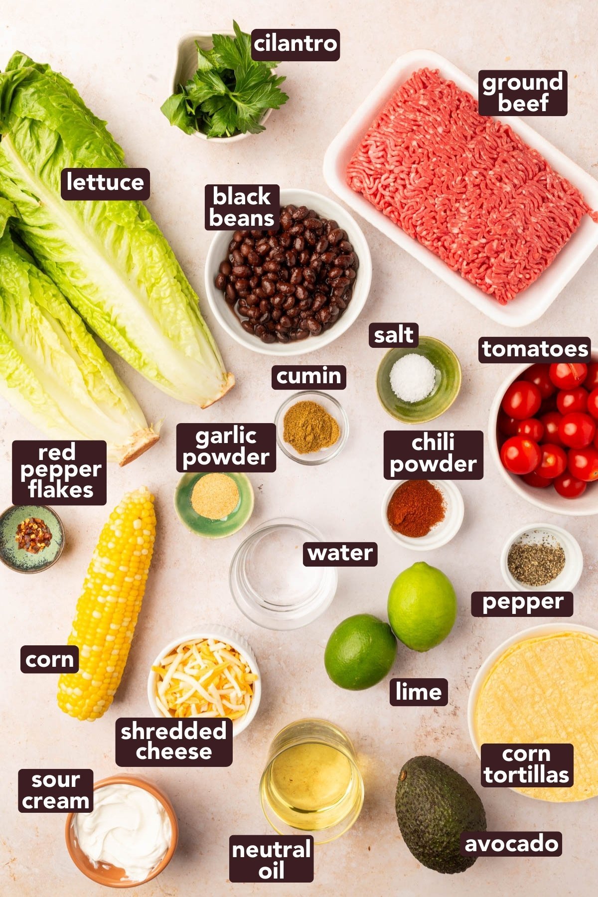Flat lay of fresh taco salad ingredients including romaine lettuce, ground beef, corn tortillas, avocado, tomatoes, black beans, cheese, and spices.